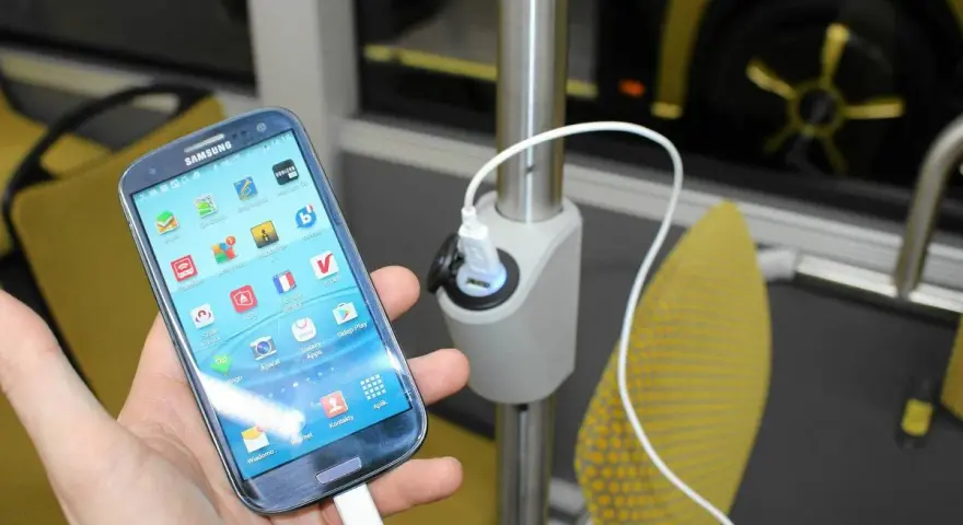 Dłoń trzyma smartfon podłączony do ładowarki USB zamontowanej na słupku w autokarze.