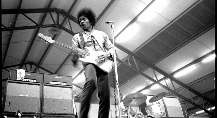 Jimi Hendrix, icône du jimi hendrix style de musique, électrise la scène avec sa guitare blanche. Amplis Marshall et batterie en arrière-plan.