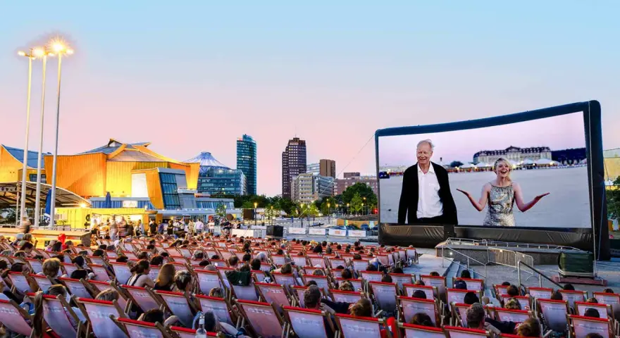 Ein lauer Sommerabend im Freiluftkino Charlottenburg. Menschen sitzen in Liegestühlen und schauen auf eine Leinwand, auf der ein Film mit einem älteren Mann und einer jungen Frau läuft. Im Hintergrund die Berliner Skyline.