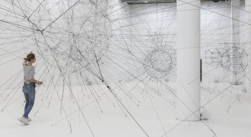 Une femme admire une installation de Tomas Saraceno, un réseau complexe de fils noirs tendus dans un espace blanc.