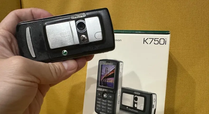 Dłoń trzyma czarny telefon Sony Ericsson K750i, obok stoi pudełko z tym modelem.