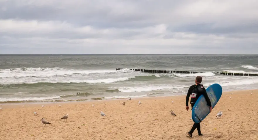 Surfer idzie plażą w Juracie, szukając fal. To jedna z wielu atrakcji tego miejsca.