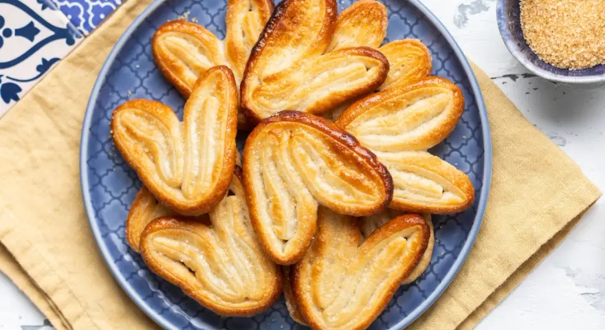 Un piatto blu pieno di dolci con pasta sfoglia dorati e croccanti, cosparsi di zucchero.
