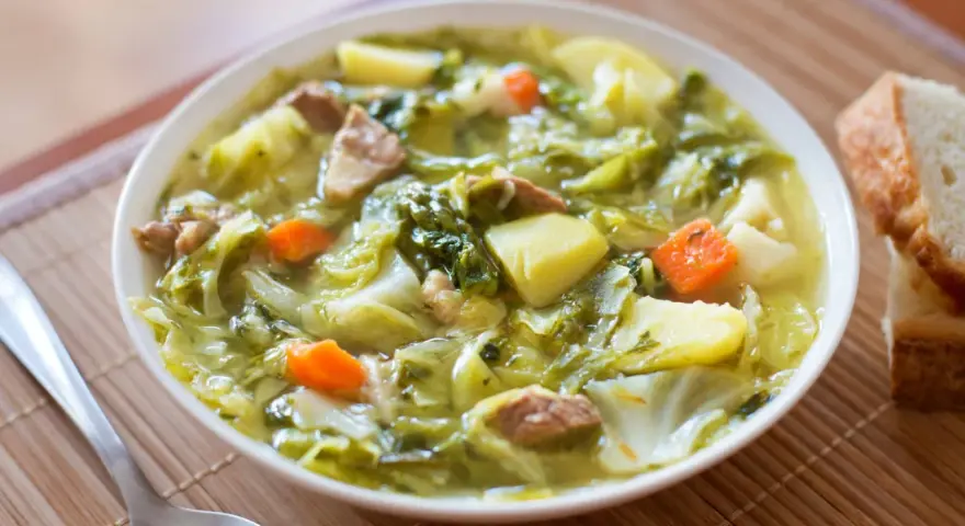 Un bol de soupe aux choux réconfortante, avec des morceaux de chou, pommes de terre, carottes et viande. Du pain frais à côté.