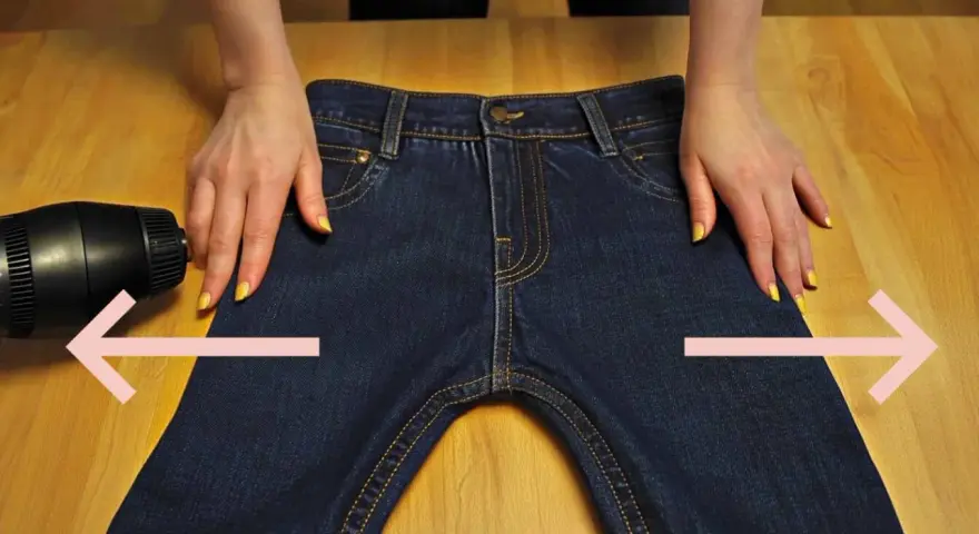 Manos sujetan pantalones vaqueros oscuros sobre mesa de madera. Secador de pelo apunta a la tela. Aprende como estirar pantalones vaqueros.