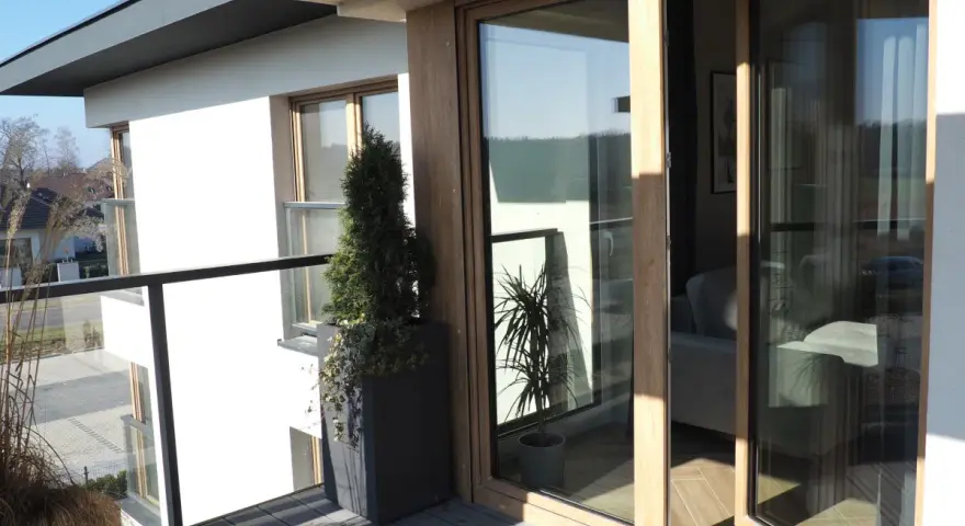 Nowoczesny balkon z drewnianymi, podwójnymi drzwiami balkonowymi. Okna dzielone poziomo wpuszczają dużo światła.