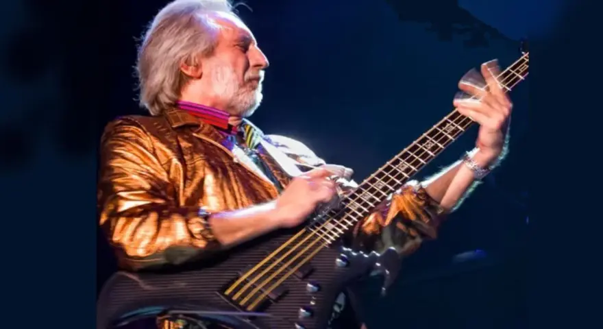 Ein Mann mit grauem Haar und Bart spielt Bassgitarre. Er trägt eine glänzende goldene Jacke und bunte Krawatte. Er ist der beste Bassist der Welt.