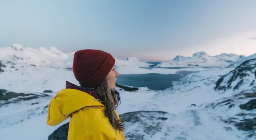 Kvinna i gul jacka och röd mössa njuter av utsikten över snöklädda berg och hav. Perfekt för sportlov i Norge.
