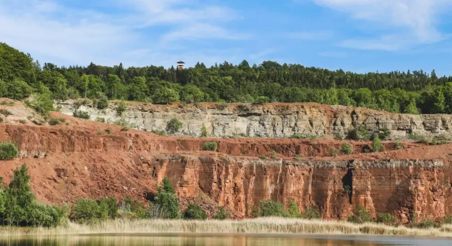 En sjö speglar Sveriges Grand Canyon, en imponerande klippformation med lager av röd och grå sten, krönt av en grönskande skog.