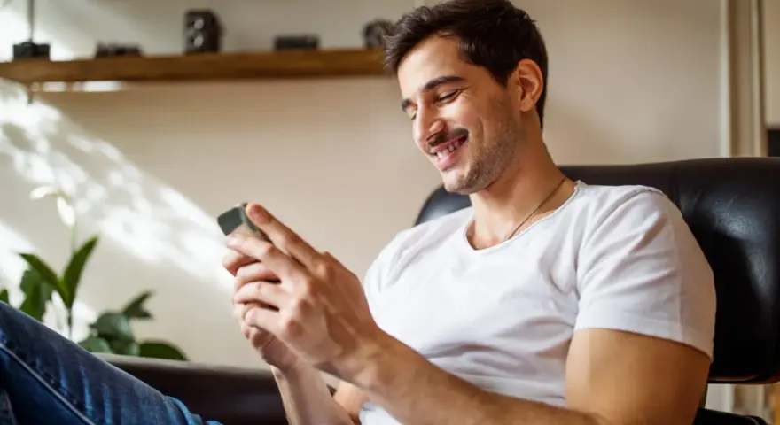Uśmiechnięty mężczyzna w białej koszulce siedzi w fotelu i patrzy na telefon.