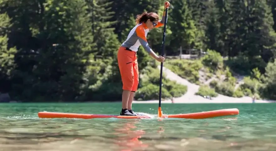 Kobieta w pomarańczowych spodenkach i sportowej bluzce uprawia pływanie na supie po spokojnym jeziorze. W tle zielone drzewa i jasna plaża.