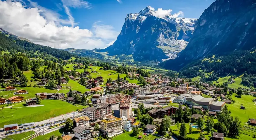 A scenic view of Grindelwald, Switzerland, nestled in a valley with lush green hills and a majestic snow-capped mountain. A train travels through the village.