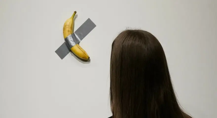 Dojrzały banan przyklejony do ściany szarą taśmą, widziany zza ramienia osoby z długimi, ciemnymi włosami.