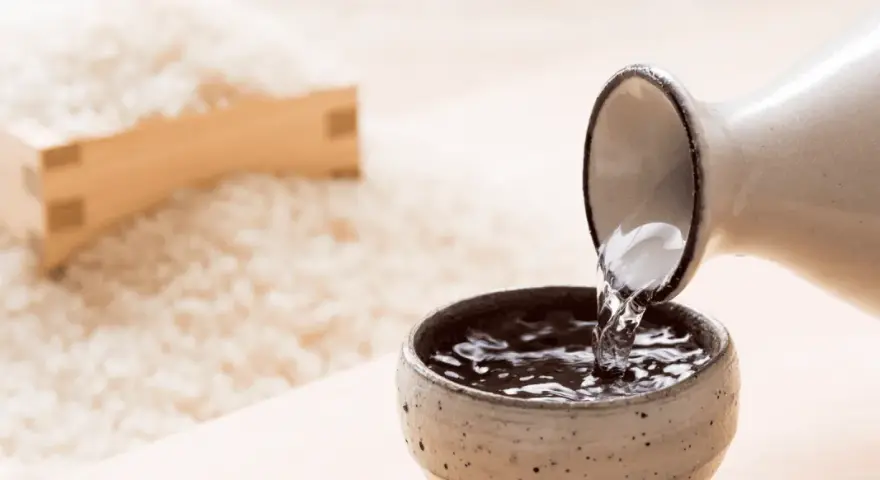 Sake przelewana z dzbanka do czarki, w tle ryż. Poznaj, jak smakuje sake.