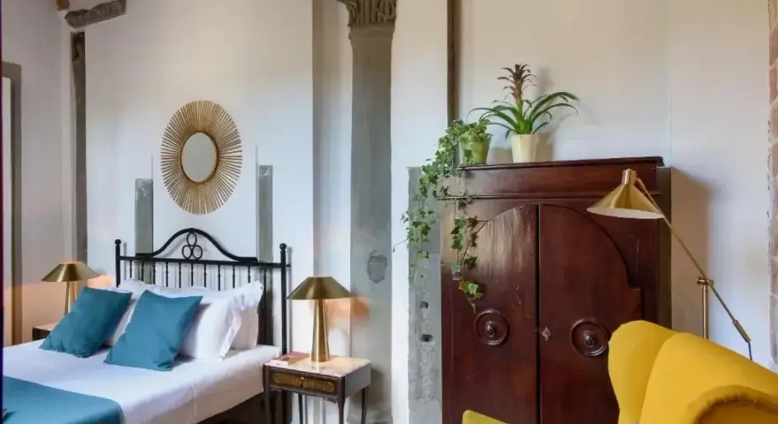 Elegancka sypialnia z łóżkiem, zabytkową szafą i żółtym fotelem. Idealne miejsce, by dowiedzieć się, gdzie nocować w Toskanii.