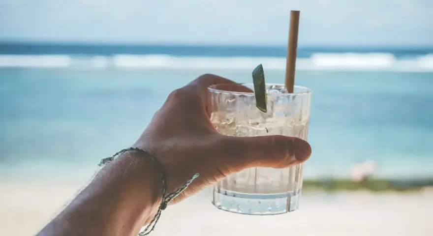 Orzeźwiający drink na plaży, z lodem i plasterkiem limonki, w dłoni z bransoletką. W tle błękitne morze i fale.