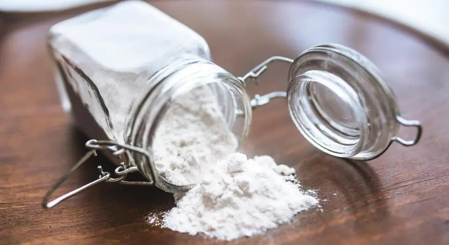 Un barattolo di vetro rovesciato su un tavolo di legno, con polvere bianca che fuoriesce, forse ingredienti per colla vinilica fatta in casa.