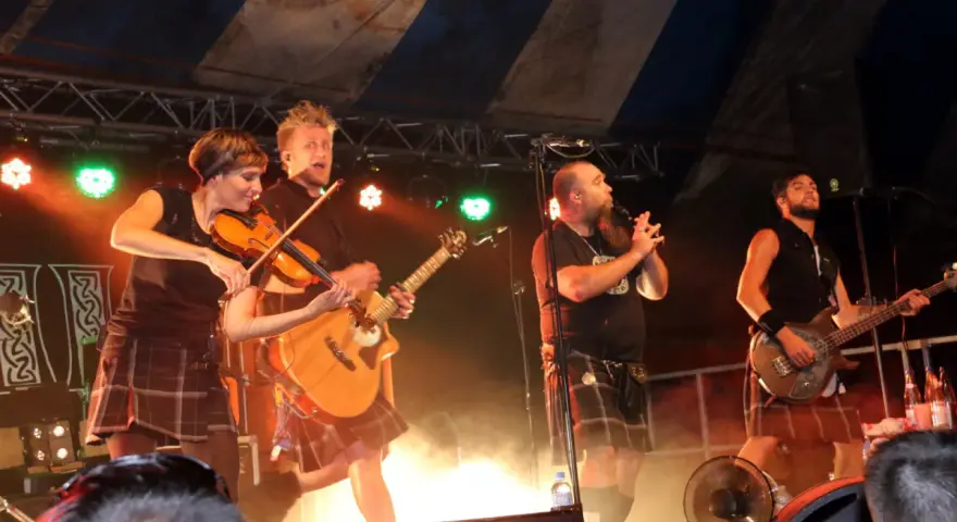 Un groupe celtique français sur scène, avec violon, guitare et basse. Les musiciens portent des kilts, créant une ambiance festive.