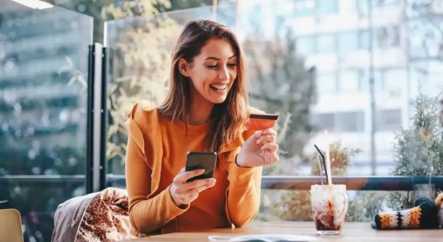 Smiling woman uses phone and credit card. She's researching if you need an LLC for a business credit card.
