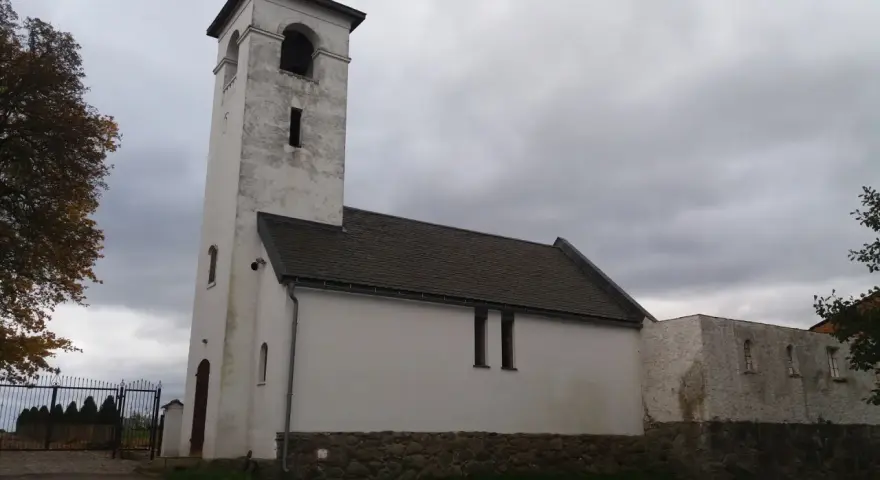 Biały, zabytkowy dwór z wieżą i dachem krytym dachówką. W oddali widać bramę i drzewa.