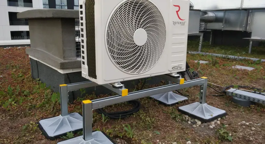Jednostka zewnętrzna klimatyzatora Rotenso na dachu, z widocznymi kanałami wentylacyjnymi i budynkiem w tle.