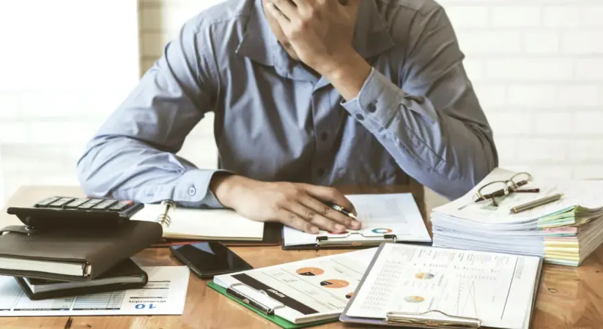 Zestaw dokumentów, kalkulator i telefon leżą na biurku. Mężczyzna z ręką na czole, przytłoczony problemami finansowymi, przegląda pismo windykacyjne.