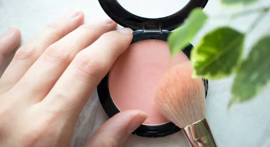 Une main applique un pinceau sur un blush poudre, révélant une douce teinte couleur blush.
