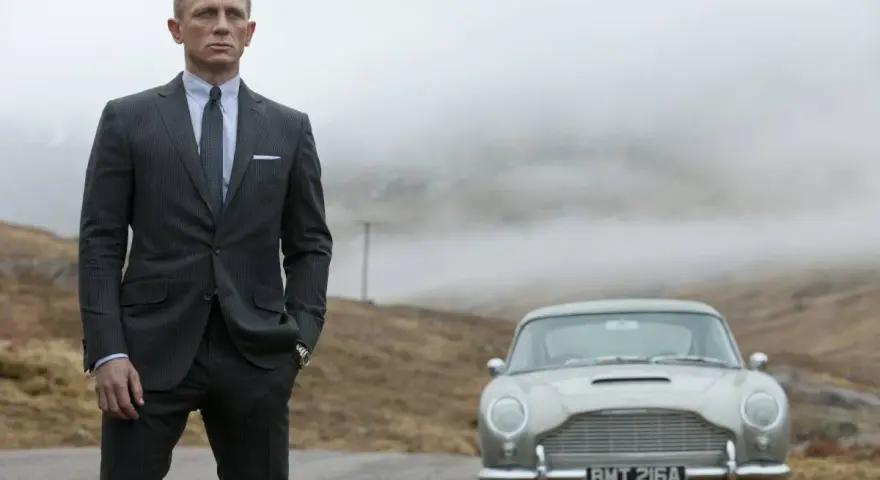 Uomo con impeccabile 007 outfit, completo gessato e cravatta, posa accanto a una Aston Martin d'epoca in un paesaggio nebbioso.