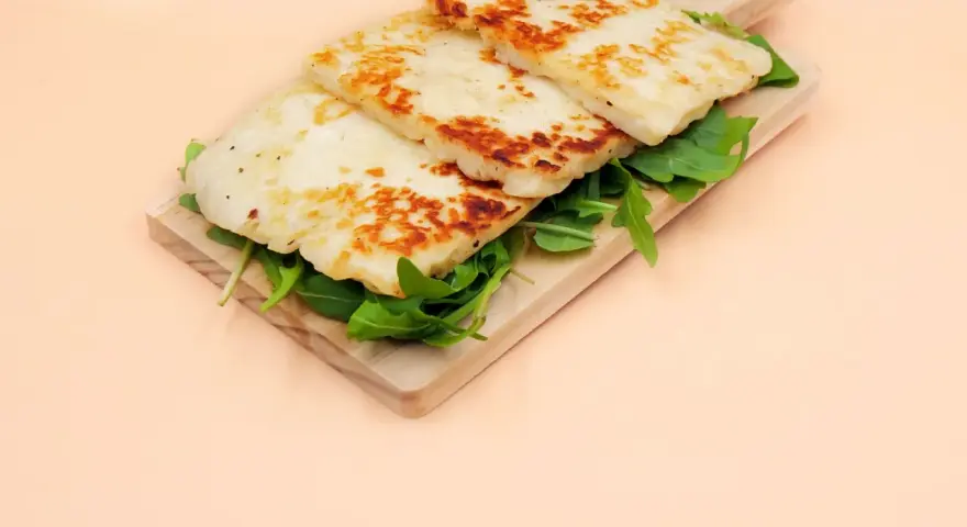 Trzy złociste plastry sera halloumi na desce z rukolą. Czy w ciąży można jeść ser halloumi? Tak, jeśli jest pasteryzowany.