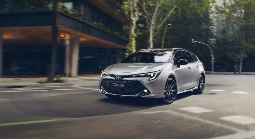 Srebrna Toyota Corolla kombi z czarnymi felgami sunie po miejskiej ulicy, otoczona zielenią i nowoczesną architekturą.