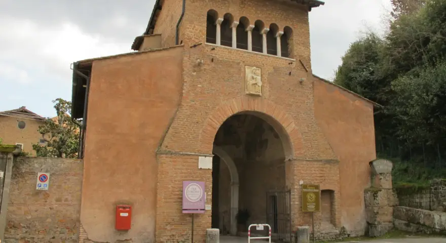 The imposing facade of Tre Fontane Abbey, with its arched entrance and upper loggia, stands against a cloudy sky.