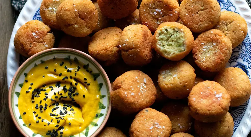 Falafel ricetta originale: croccanti palline dorate, una con un morso rivela il verde interno, accompagnate da una salsa gialla con semi di sesamo nero.