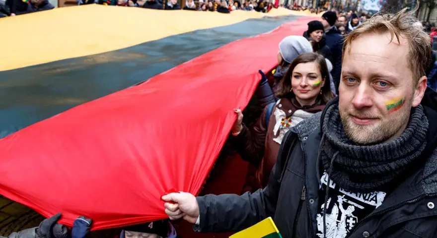 Ludzie niosą ogromną flagę Litwy podczas święta narodowego. Na twarzach uczestników widać patriotyczne barwy.