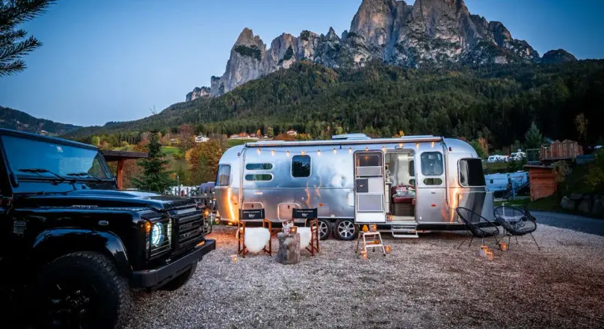 Ein Airstream-Wohnwagen und ein schwarzer Geländewagen stehen auf einem **Campingplatz Dolomiten** mit majestätischen Bergen im Hintergrund. Gemütliche Stühle und Lichter schaffen eine einladende Atmosphäre.