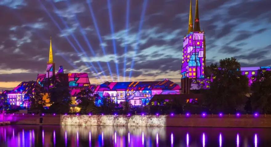 Ostrów Tumski we Wrocławiu nocą, rozświetlony kolorowymi projekcjami i niebieskimi snopami światła.