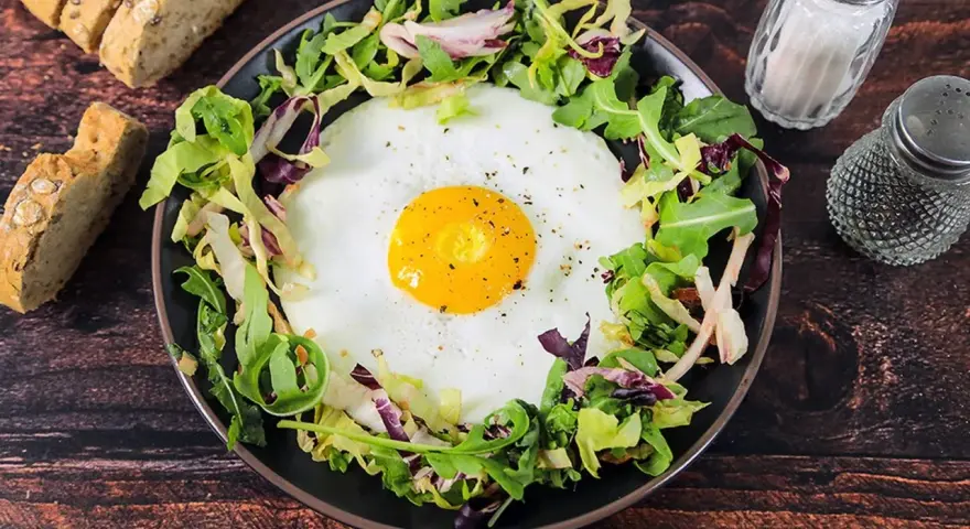 Un uovo occhio di bue su un letto di insalata mista, servito con pane integrale e condimenti.