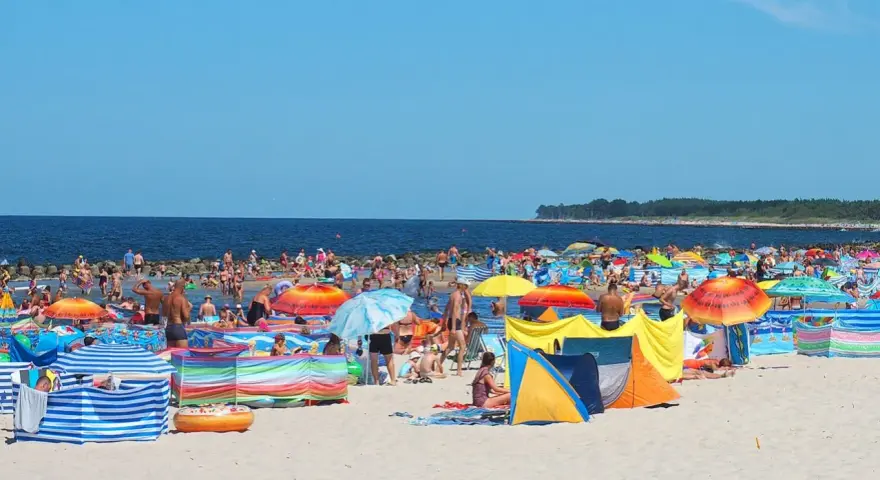 Tłum ludzi na plaży w Darłówku Wschodnim, kolorowe parawany i namioty chronią przed słońcem.