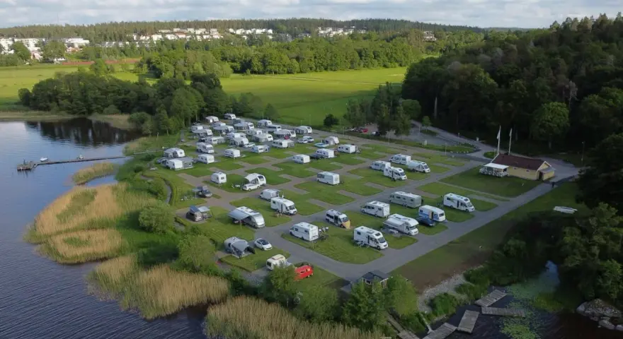 Full campingplats med husbilar och husvagnar vid en sjö, med grönskande träd och bebyggelse i bakgrunden. En idyllisk plats för camping i Alingsås.