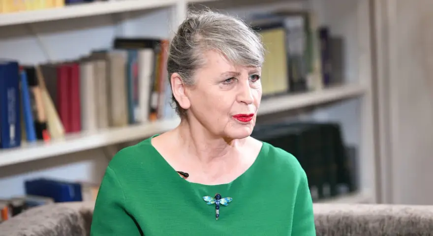 Joanna Wnuk-Nazarowa w zielonej bluzce z broszką w kształcie ważki, na tle regałów z książkami.