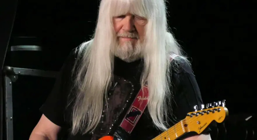 Andy Scott mit seinen charakteristischen langen, weißen Haaren spielt auf einer orangen E-Gitarre.