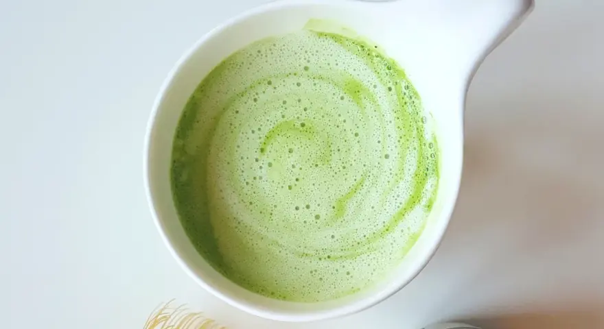 Zielony, spieniony napój matcha w białej miseczce, obok bambusowa miotełka i puszka z proszkiem. Idealne co na pobudzenie zamiast kawy.