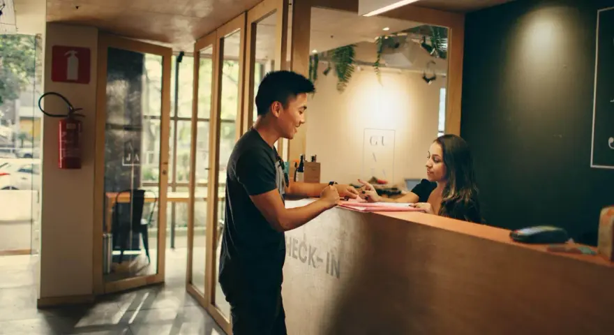 Mężczyzna przy ladzie check-in pyta hotelarkę, ile zarabia. W tle widać drzwi i gaśnicę.