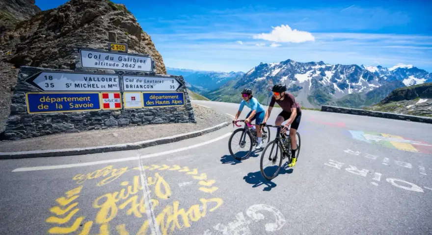 Ciclisti in salita verso il Col du Galibier, con montagne innevate sullo sfondo e segnaletica stradale.