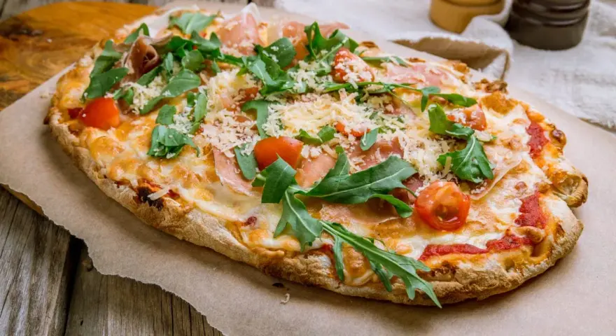 Pinsa co to? Aromatyczna, podłużna pizza z rukolą, pomidorkami i szynką parmeńską, posypana parmezanem.