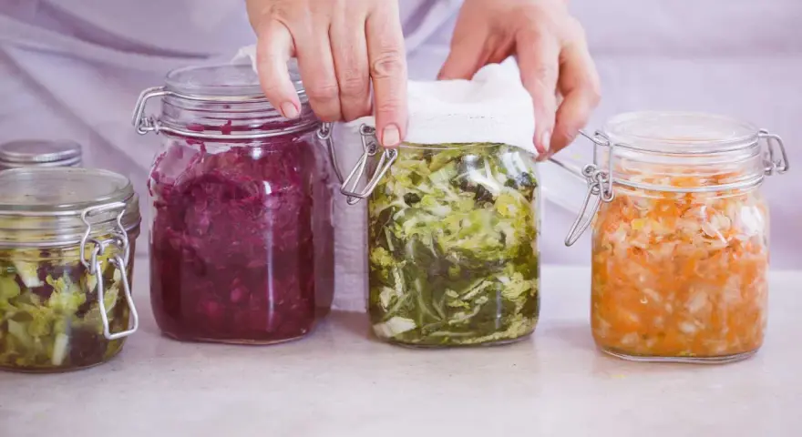 Hände bedecken ein Glas mit fermentiertem Gemüse, Teil einer Auswahl an probiotischen Lebensmitteln in Gläsern.
