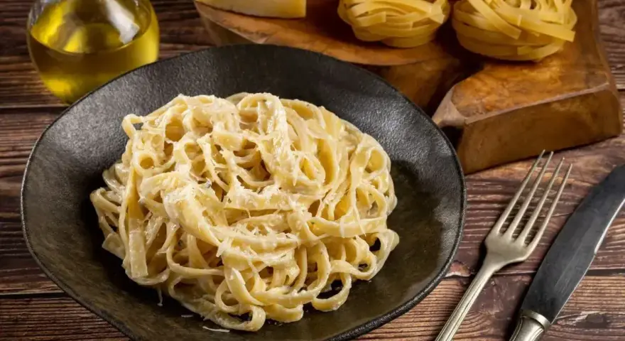 Pyszne Fettuccine Alfredo przepis na talerzu, obok surowy makaron, ser i oliwa. Idealne danie dla miłośników włoskiej kuchni.