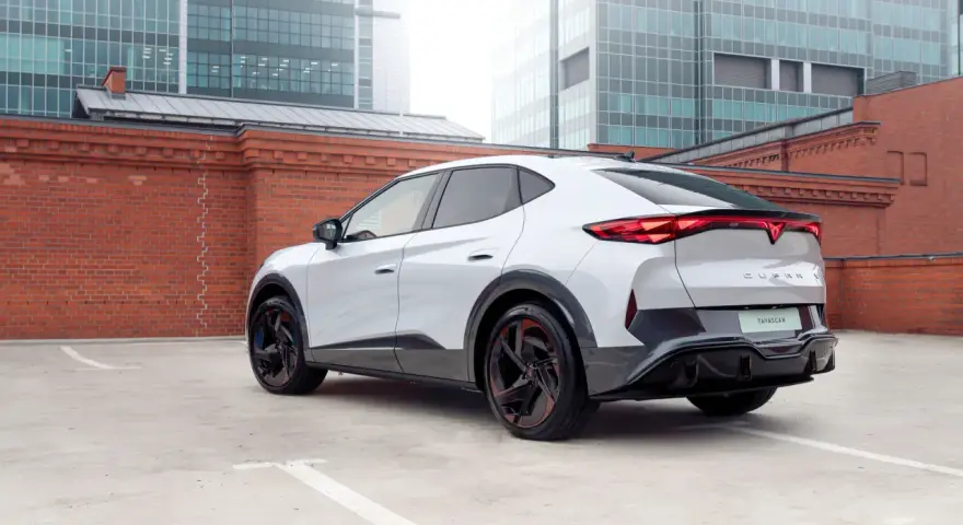 Biały elektryczny SUV Cupra Tavascan z czerwonymi akcentami na felgach, zaparkowany na tle ceglanego muru i nowoczesnych budynków.