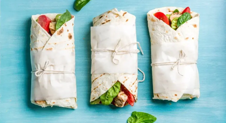 Trzy keto tortille z siemienia lnianego, zawinięte w papier i przewiązane sznurkiem, z widocznym nadzieniem z kurczaka, pomidorów i szpinaku.