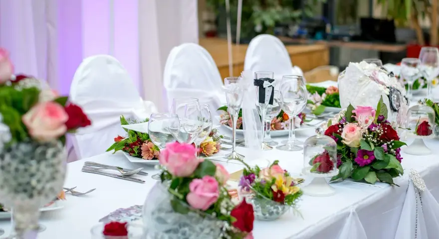 Elegancki, samodzielny wystrój sali weselnej: białe pokrowce na krzesła, bukiety róż i goździków, kieliszki do wina i szampana.