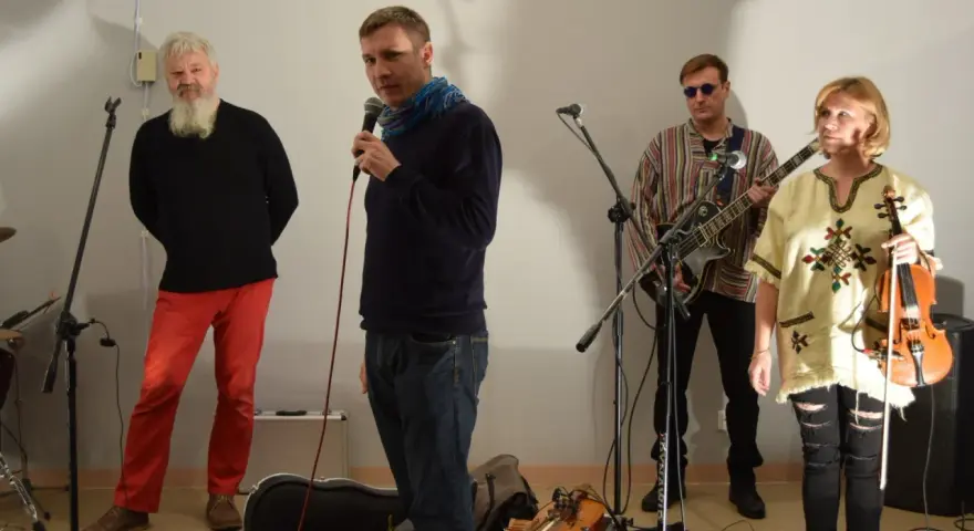 Orkiestra Świętego Mikołaja przygotowuje się do występu. Wokalista z mikrofonem, gitarzysta, skrzypaczka i brodaty mężczyzna w czerwonych spodniach.